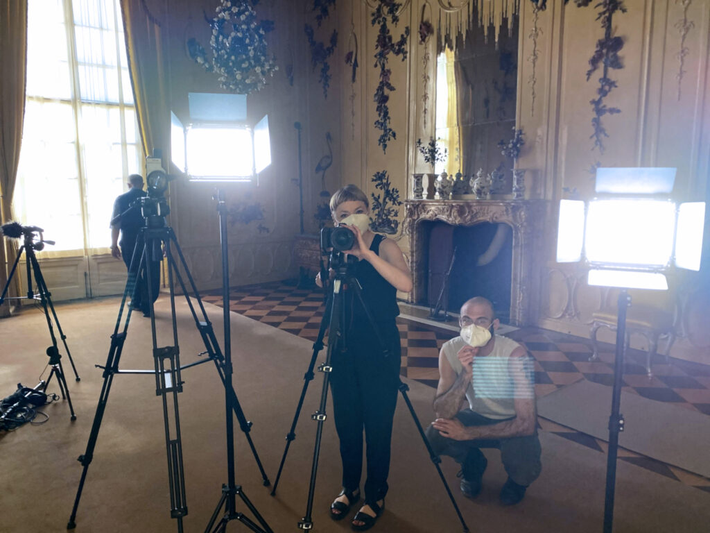 The filmmaker stands behind a camera beside lights and other tripods in the ornately carved, yellow room of the Sans Souci Palace. The sound designer squats beside her. Both are masked and look into the lens.